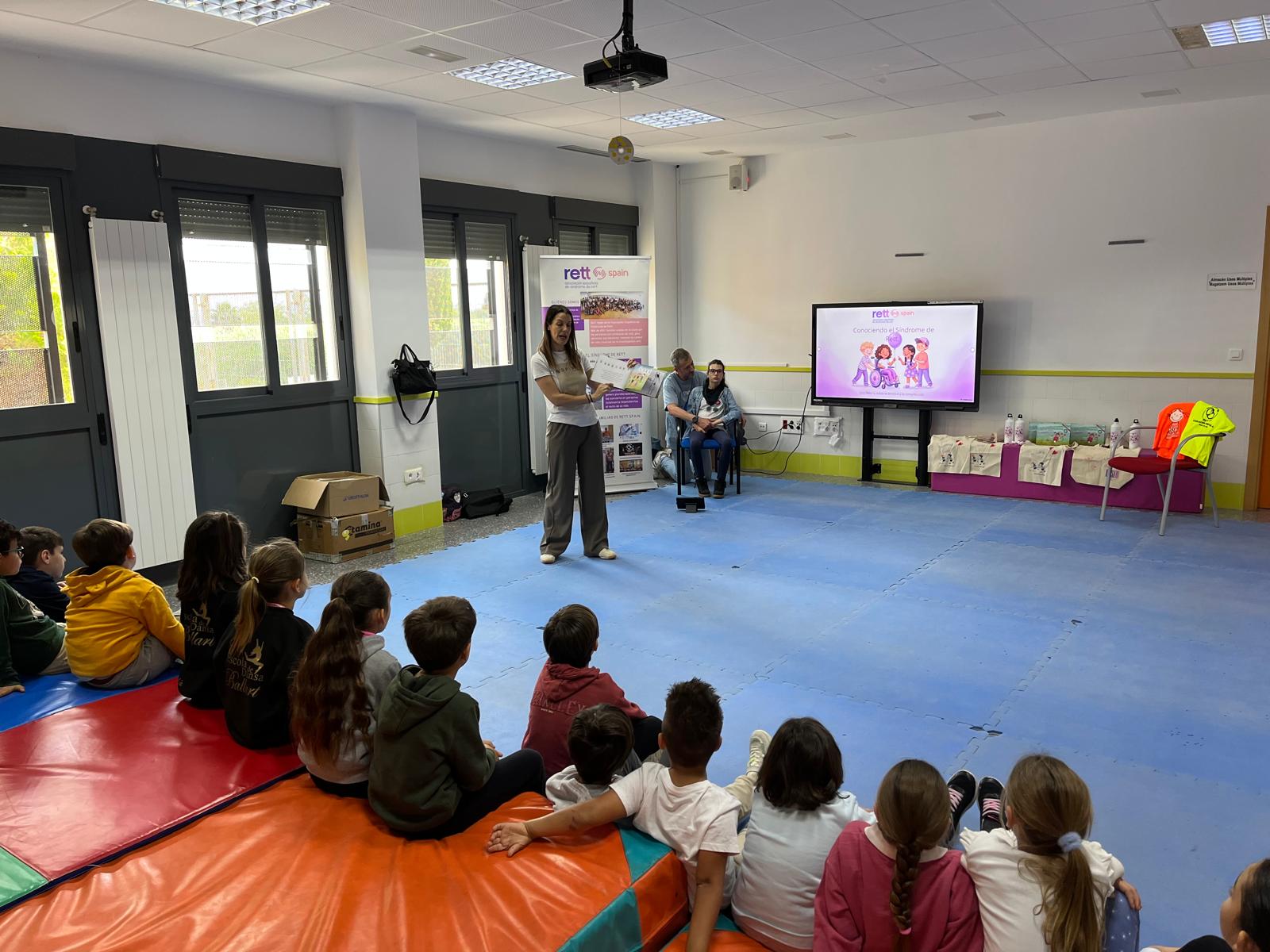 Mujer joven vestida con pantalón oscuro y camiseta blanca de manga corta está de pie mientras sujeta el libro abierto de La princesa sin palabras, detrás sentados están un padre Rett y su hija Rett junto a un roll up de Rett Spain. Varios niños sentados en el suelo atienden a la profesora