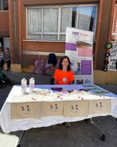 mesa con productos solidario de Rett Spain, hay una chiba sentada con camiseta naranja con dibujo de niña con coletas y detrás se ve el Roll Up de Rett Spain